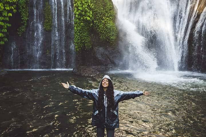 Young happy woman in raincoat enjoying waterfall in Bali