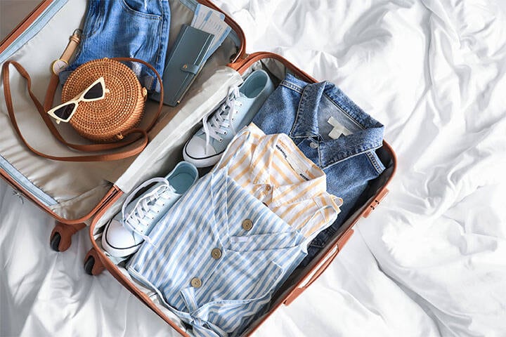 Open packed Suitcase on bed, with linen shirts, sneakers, woven cross-body bag, sunglasses and a travel wallet