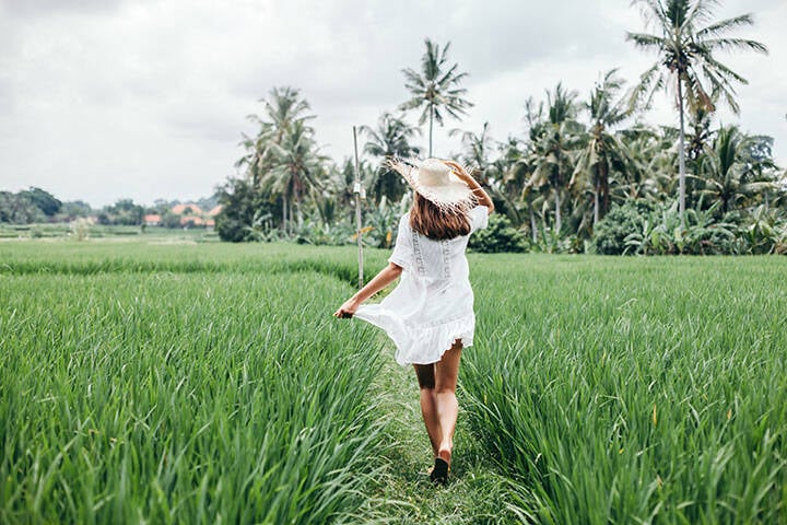 Young woman in a short-sleeve loose  white dress, sandals and wide-brim hat, walking in rice field in Bali