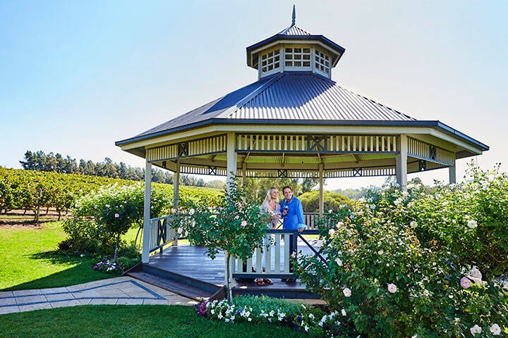 A couple stands under a gazebo surrounded by lush green bushes and vibrant flowers at Sittella Winery & Restaurant in Herne Hill. 