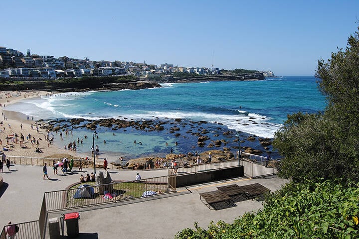 A vibrant image of Bronte Bogey Hole in Sydney, featuring clear blue water surrounded by rocky cliffs. People can be seen enjoying a swim in the natural rock pool under the warm sun, with lush greenery in the background creating a relaxing beach atmosphere.