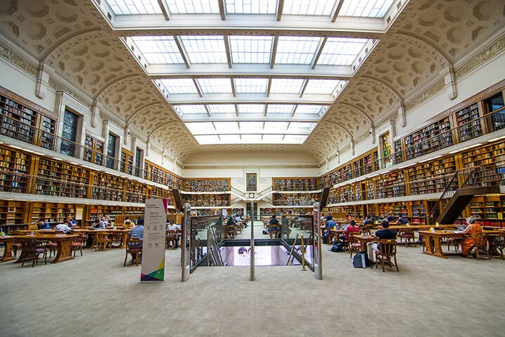 An interior view of the State Library of New South Wales in Sydney, showcasing books lining the walls. People are seated at desks, engrossed in reading, creating a quiet and scholarly atmosphere within the beautifully designed space.