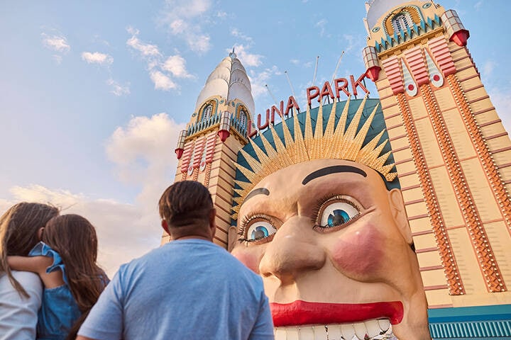 A family stands in front of the entrance to Luna Park in Sydney, gazing up at the iconic entryway figure against a bright blue sky.