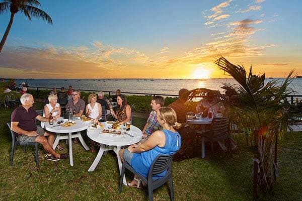 Guests sitting outside on grass next to barbecue and drinks overlooking sunset water view at Darwin Trailer Boat Club, Darwin.