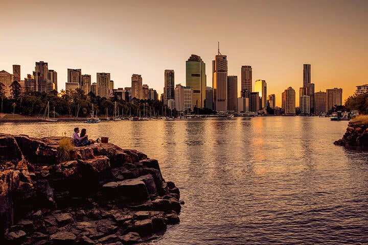 A couple having a picnic by the Brisbane River on a rock at Kangaroo Point while gazing at Brisbane’s River Side Centre and the sunset 