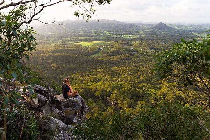 A young man sits, quietly meditating on top of a mountain with expansive views near Noosa Heads, Australia.