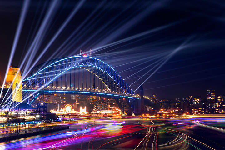 Sydney Harbour Bridge and bay at night with lights beaming from the top for Vivid Festival, and boat lights in the bay. 