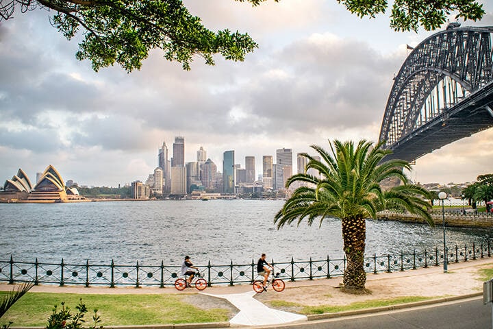 Wide Shot of Tourists Biking along Sydney Harbour, with a view of the Sydney Skyline, Milsons Point, Waterfront, Sydney Harbour Bridge, and Opera House on a Summer Evening