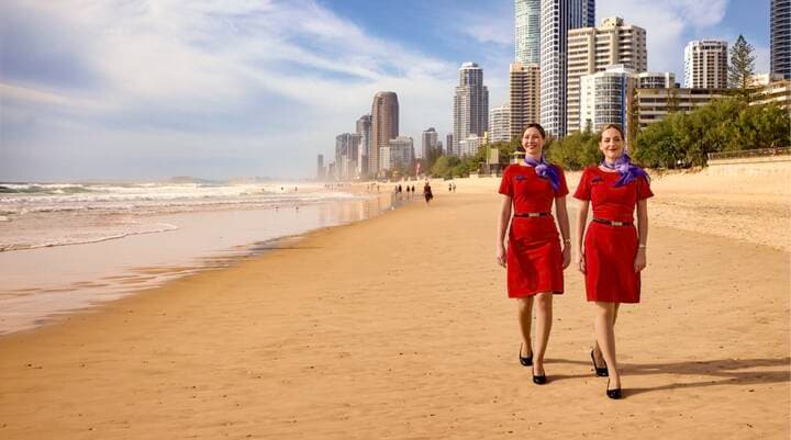 A Virgin Australia staff member in a red uniform smiles while assisting a guest in a purple blazer at a self-service check-in area, holding a mobile device. Behind them are airport kiosks and a screen displaying “Priority Bag drop.”