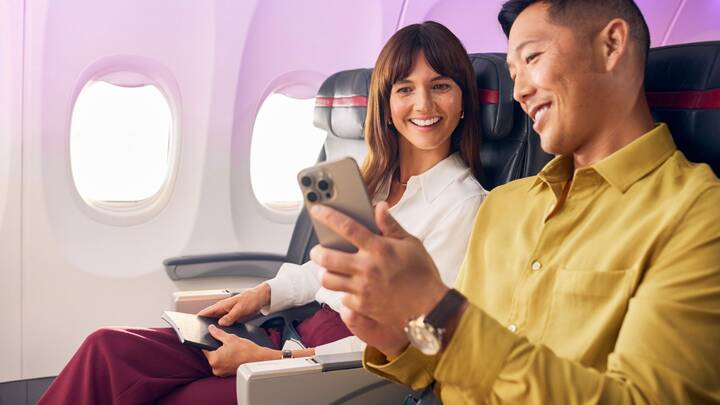 A Virgin Australia staff member in a red uniform smiles while assisting a guest in a purple blazer at a self-service check-in area, holding a mobile device. Behind them are airport kiosks and a screen displaying “Priority Bag drop.”