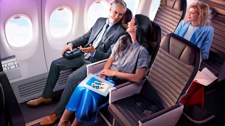 Three passengers seated in a Virgin Australia Boeing 737 Economy X cabin, showcasing the extra legroom. In the foreground, a man in a grey suit and a woman in a blue skirt and patterned shirt sit in the exit row, smiling and talking. In the background, a woman in a blue blazer is seated in a standard Economy X row. The seats are dark grey with purple accents and "Economy X" branding on the headrests.