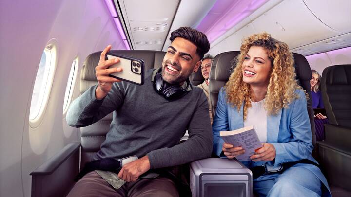 Two smiling passengers seated on an aircraft take a selfie with a smartphone, with other travellers visible behind them in a softly lit cabin.