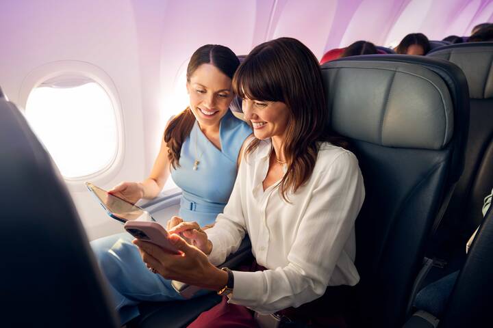 A Virgin Australia staff member in a red uniform smiles while assisting a guest in a purple blazer at a self-service check-in area, holding a mobile device. Behind them are airport kiosks and a screen displaying “Priority Bag drop.”