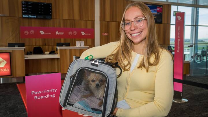 A Virgin Australia staff member in a red uniform smiles while assisting a guest in a purple blazer at a self-service check-in area, holding a mobile device. Behind them are airport kiosks and a screen displaying “Priority Bag drop.”