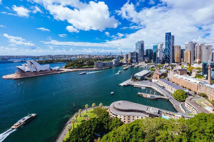 Aerial view of Sydney Harbour with the Opera House, cruise ships docked at Circular Quay, and city skyline, representing Virgin Australia’s relaunch of check-in and bag drop service for cruise passengers.