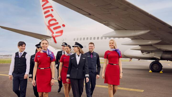 A Virgin Australia staff member in a red uniform smiles while assisting a guest in a purple blazer at a self-service check-in area, holding a mobile device. Behind them are airport kiosks and a screen displaying “Priority Bag drop.”