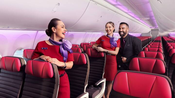 Three Virgin Australia cabin crew members stand in the aisle of an airplane, smiling and chatting. Two women wear red uniforms with purple scarves, and one man wears a black uniform. Rows of red-and-black seats line both sides of the cabin, with soft purple lighting along the ceiling.