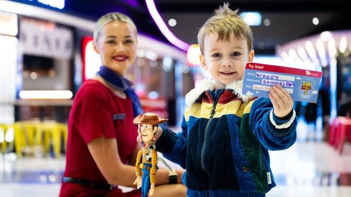 A Virgin Australia staff member in a red uniform smiles while assisting a guest in a purple blazer at a self-service check-in area, holding a mobile device. Behind them are airport kiosks and a screen displaying “Priority Bag drop.”