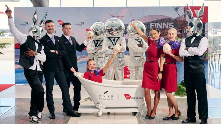 A Virgin Australia staff member in a red uniform smiles while assisting a guest in a purple blazer at a self-service check-in area, holding a mobile device. Behind them are airport kiosks and a screen displaying “Priority Bag drop.”