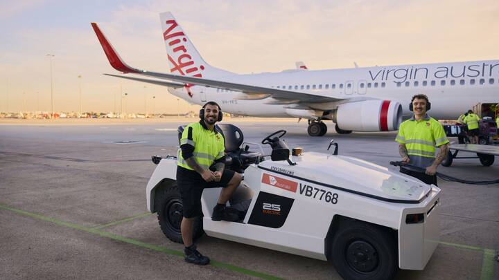 A Virgin Australia staff member in a red uniform smiles while assisting a guest in a purple blazer at a self-service check-in area, holding a mobile device. Behind them are airport kiosks and a screen displaying “Priority Bag drop.”
