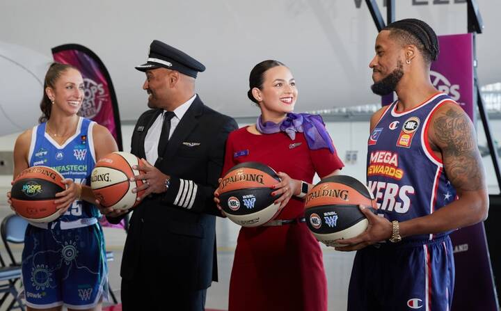 A Virgin Australia staff member in a red uniform smiles while assisting a guest in a purple blazer at a self-service check-in area, holding a mobile device. Behind them are airport kiosks and a screen displaying “Priority Bag drop.”