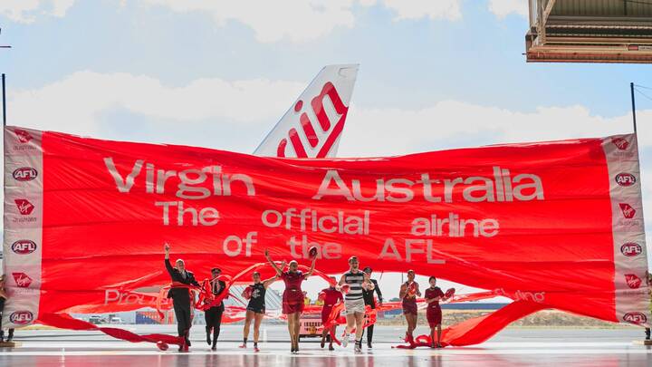 A group of Virgin Australia cabin crew, pilots, and AFL players in uniform running through a large red banner inside an airport hangar to reveal the tail of a Virgin Australia aircraft. The banner reads "Virgin Australia: The official airline of the AFL" in white text.