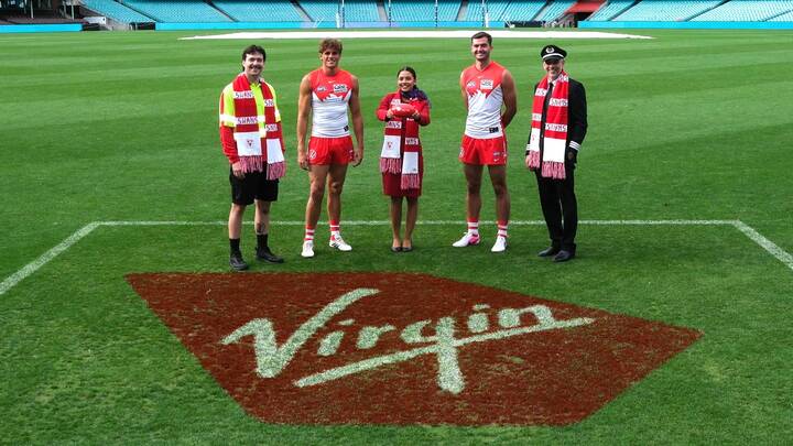 Five people stand on a football field at a stadium in front of a large Virgin logo painted on the grass, including two Sydney Swans AFL players in uniform, two Virgin Australia cabin crew in red uniforms, and another person wearing a Sydney Swans scarf, posing for a promotional photo.
