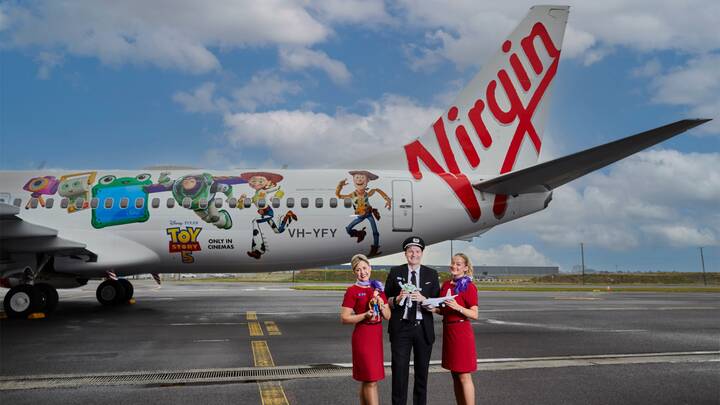 A Virgin Australia staff member in a red uniform smiles while assisting a guest in a purple blazer at a self-service check-in area, holding a mobile device. Behind them are airport kiosks and a screen displaying “Priority Bag drop.”