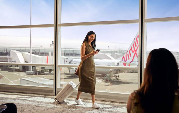 A Virgin Australia staff member in a red uniform smiles while assisting a guest in a purple blazer at a self-service check-in area, holding a mobile device. Behind them are airport kiosks and a screen displaying “Priority Bag drop.”