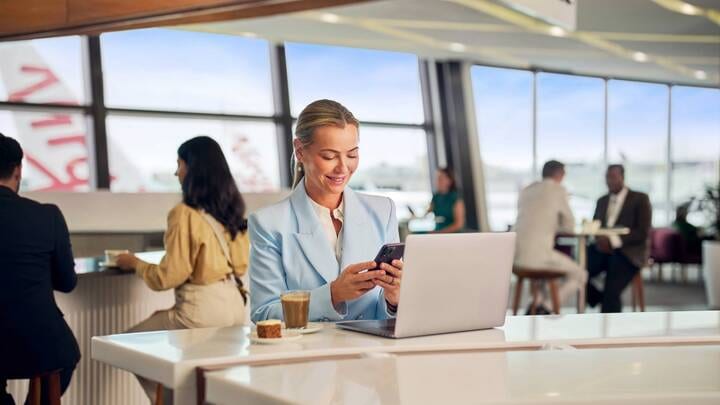 A smiling woman stands in an airport terminal looking out a large window at a Virgin Australia aircraft parked on the tarmac.